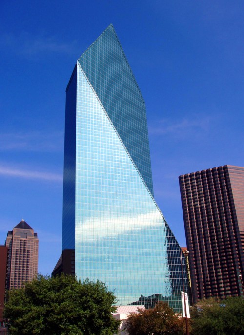 A tall, modern glass skyscraper with a distinctive triangular shape against a blue sky, surrounded by other buildings.