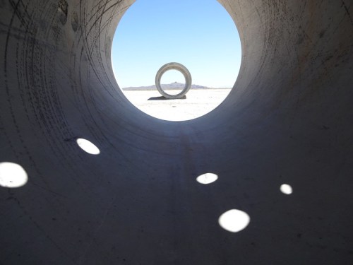 View through a concrete tunnel, framing a circular sculpture against a clear blue sky and distant landscape.