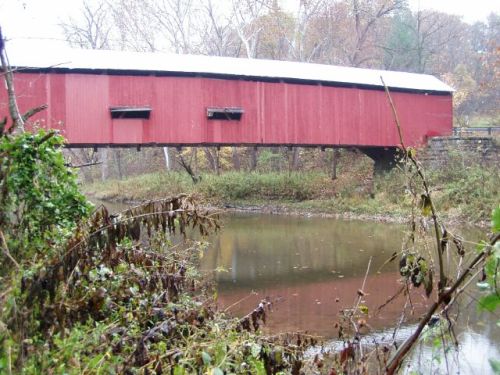 A red covered bridge spans a calm river, surrounded by autumn foliage and overgrown vegetation.