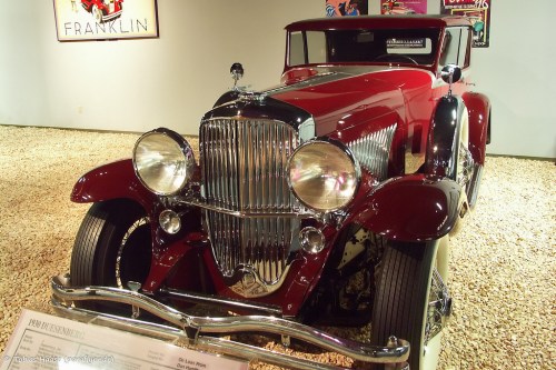 A vintage red car with chrome details displayed in a museum setting, featuring a shiny grille and large headlights.
