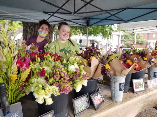 Two smiling vendors stand behind a flower stall, showcasing vibrant bouquets under a tent at a market.