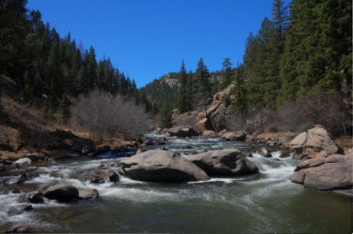 A serene river flows through a rocky landscape, surrounded by lush green trees under a clear blue sky.