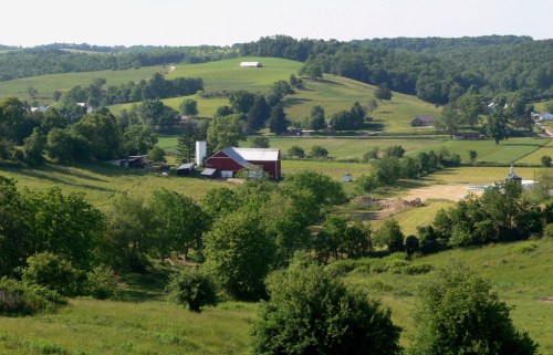 A scenic view of rolling green hills with a red barn and farm buildings nestled among trees and fields.