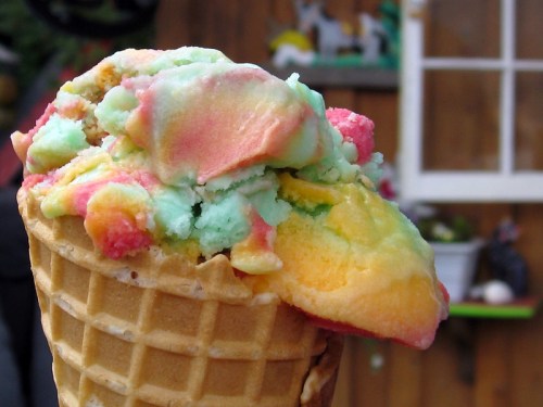 A colorful scoop of rainbow sherbet ice cream in a waffle cone, set against a rustic wooden background.