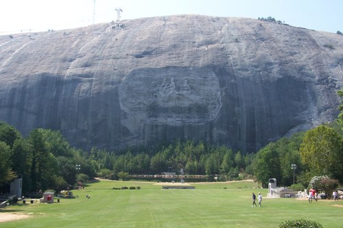 A large granite mountain with a carved relief, surrounded by green trees and a grassy area with people walking.