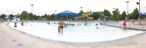 A panoramic view of a busy outdoor pool with people swimming, lounging, and a water slide in the background.