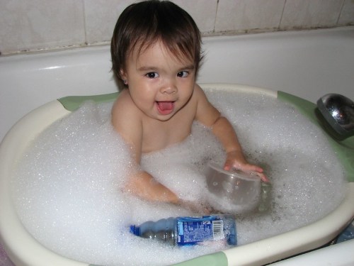 A smiling toddler playing in a bubble-filled bathtub, holding a cup and a water bottle.
