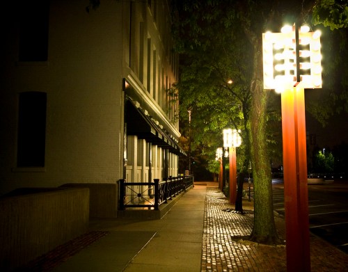 A quiet, illuminated sidewalk at night, lined with glowing street lamps and trees, beside a brick building.