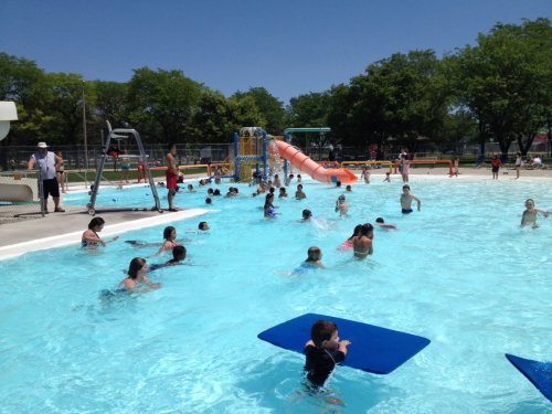 A busy outdoor pool filled with children swimming and playing, featuring slides and a sunny blue sky.