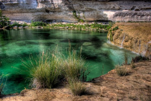 A serene green lake surrounded by rocky cliffs and lush grass, reflecting the natural beauty of the landscape.