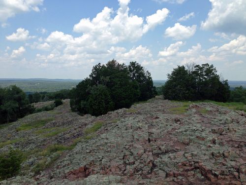 A rocky landscape with scattered trees under a partly cloudy sky, overlooking a distant green valley.
