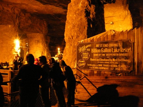 A dimly lit cave entrance with a sign for Billion Gallon Lake Resort and people gathered near a wooden fence.