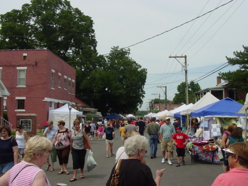 A bustling street market with people browsing tents and stalls, surrounded by trees and buildings on a sunny day.