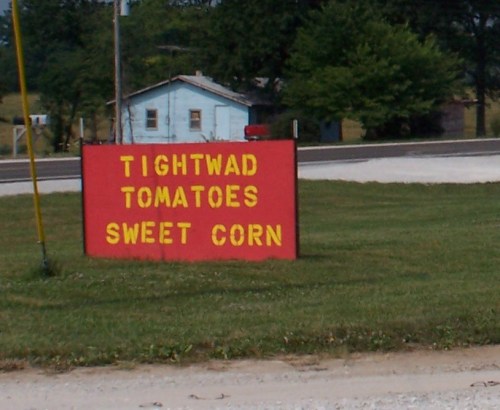 Sign advertising "Tightwad Tomatoes" and "Sweet Corn" on a grassy area near a road.