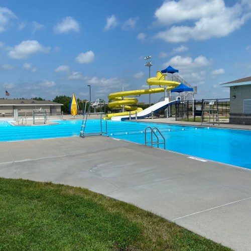 A bright outdoor swimming pool with a water slide, lounge chairs, and a clear blue sky in the background.