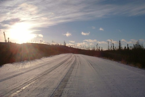 A snowy road stretches into the distance under a bright sky, with trees lining the sides and the sun setting in the background.