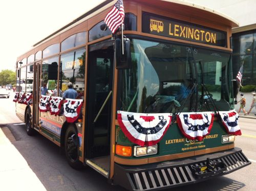A decorated trolley bus with patriotic bunting, labeled "Lexington," parked on a city street during a sunny day.