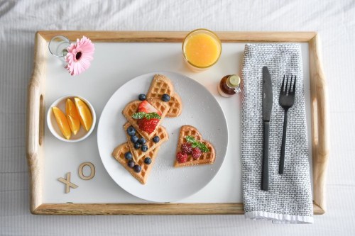 A breakfast tray with heart-shaped waffles, fresh fruit, orange juice, and a flower, set on a light background.