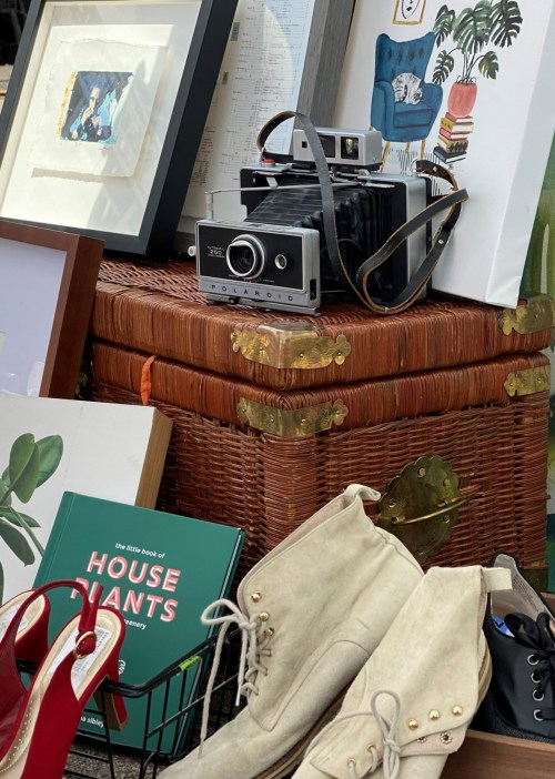 A vintage Polaroid camera sits atop woven baskets, surrounded by framed art, books, and stylish footwear.