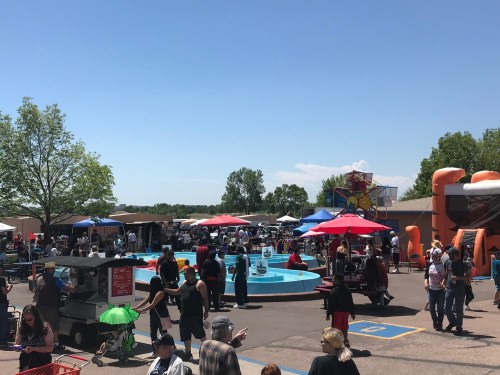 A busy outdoor event with people enjoying a pool, food stalls, and colorful umbrellas under a clear blue sky.