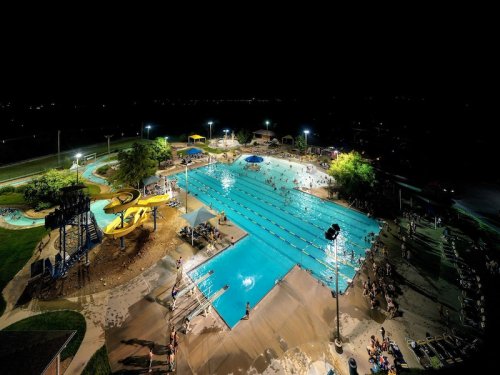 Aerial view of a brightly lit outdoor swimming pool at night, with water slides and people enjoying the water.
