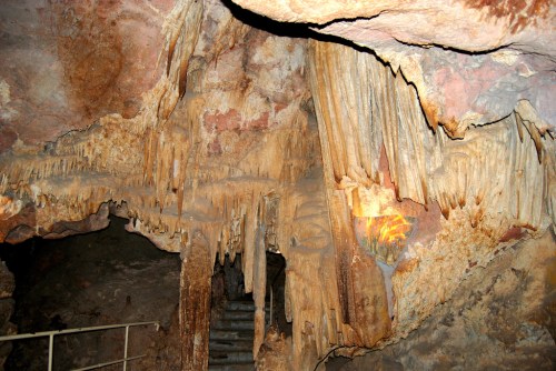Stalactites and stalagmites in a cave, with textured rock formations and a staircase in the background.