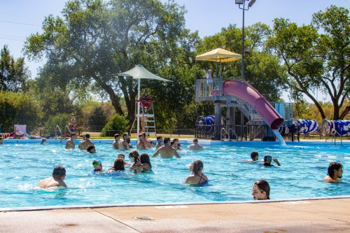 A busy outdoor pool with people swimming, a water slide, and a lifeguard on duty under a clear blue sky.