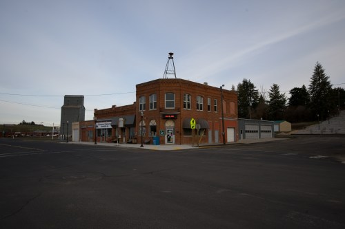 Historic brick building on a street corner, featuring a clock tower and nearby shops, with a clear sky above.