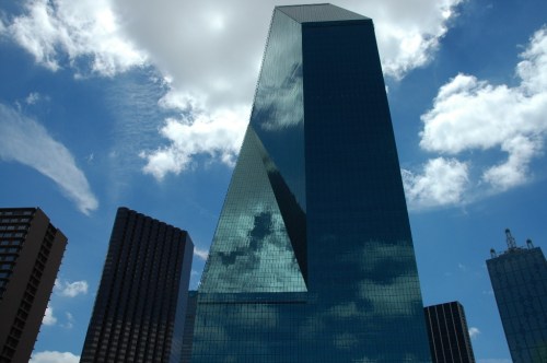 A tall, modern glass building reflecting clouds and blue sky, surrounded by other skyscrapers.