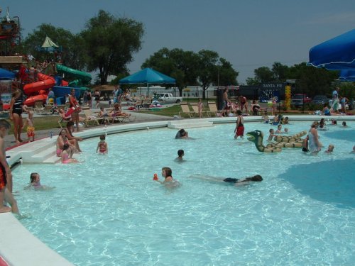 A busy pool scene with children swimming, playing, and lounging on floats, surrounded by umbrellas and water slides.
