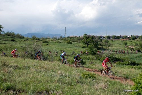 A group of mountain bikers racing through a grassy trail with hills and clouds in the background.