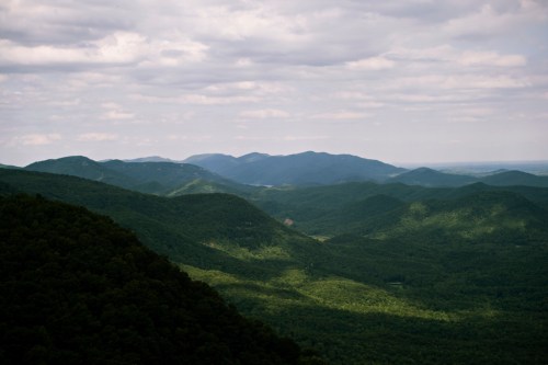 A panoramic view of rolling green mountains under a cloudy sky, with varying shades of light and shadow across the landscape.