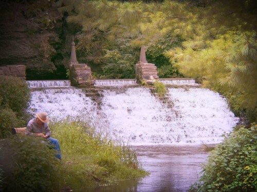 A serene scene of a waterfall cascading over stones, with a person sitting nearby, surrounded by lush greenery.