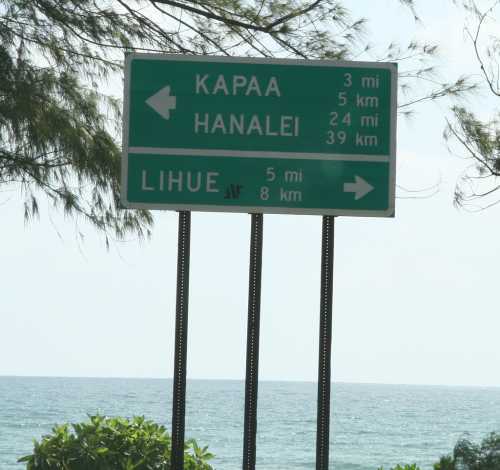 Road sign indicating directions to Kapaa (3 km), Hanalei (24 mi), and Lihue (5 mi, 8 km) with ocean in the background.