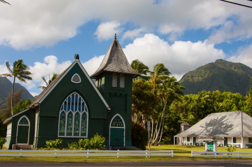 A green church with stained glass windows, surrounded by palm trees and mountains under a partly cloudy sky.
