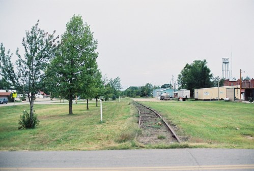A grassy area with trees alongside a set of railway tracks, leading towards a small town in the background.