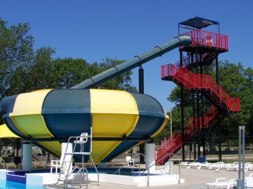 A colorful water slide with a spiral design and a tall staircase, surrounded by a pool and lounge chairs.
