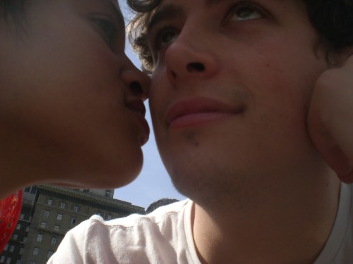 A close-up of two people, one leaning in for a kiss while the other gazes thoughtfully, with a city backdrop.