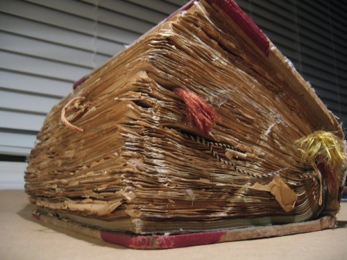 A worn, old book with frayed pages and colorful tassels, resting on a surface near a window with blinds.