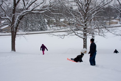 Children playing in the snow, sledding and walking in a winter landscape with trees and houses in the background.