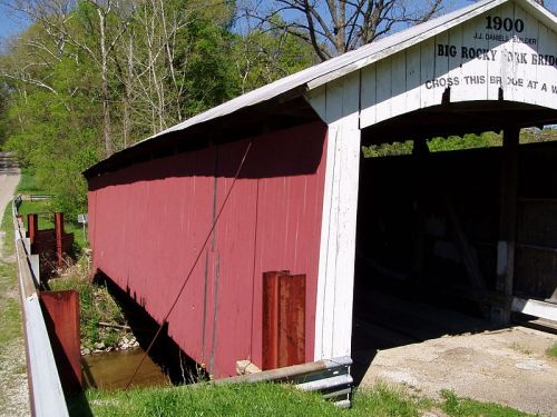 A red covered bridge with a white roof, surrounded by greenery and a gravel path, dated 1900.