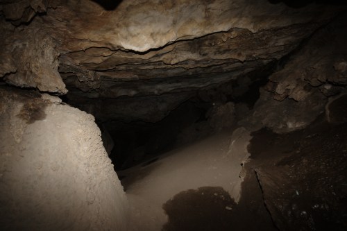 A dimly lit cave interior with rocky walls and a sandy floor, leading into a dark passageway.