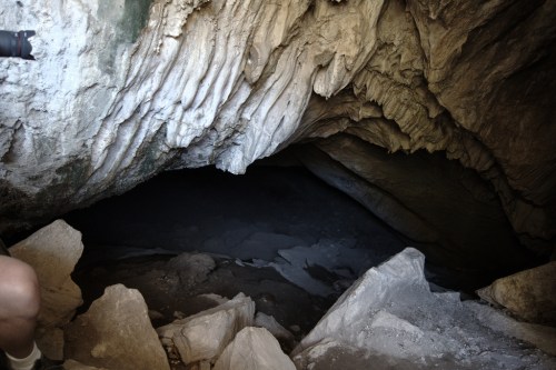 A dark cave entrance with textured rock formations and scattered stones in the foreground.