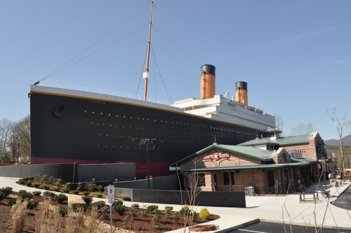 A large replica of the Titanic stands next to a brick building, set against a clear blue sky.
