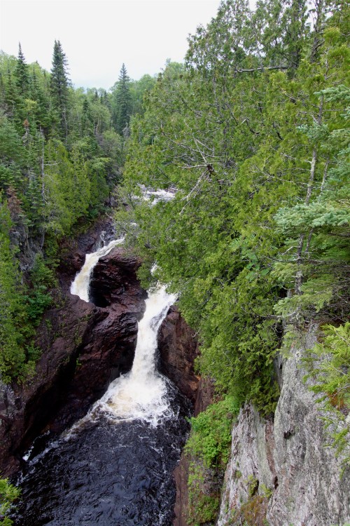 A scenic view of a waterfall cascading through rocky terrain, surrounded by lush green trees and a cloudy sky.