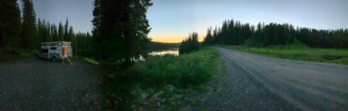 A serene lakeside scene at dusk, featuring a camper parked by the water and surrounded by tall trees and a gravel road.