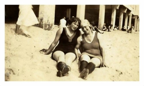 Two women in vintage swimsuits sit on the beach, smiling, with people and pillars in the background.