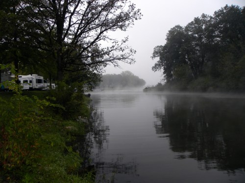 A misty river scene with trees lining the banks and a calm water surface reflecting the fog.