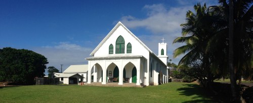 A white church with green accents, surrounded by grass and palm trees, under a blue sky with scattered clouds.