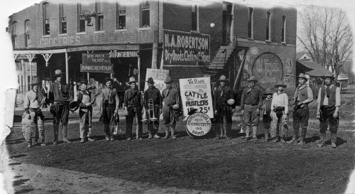 A historic black-and-white photo of a group of men in front of a building, holding instruments and a sign for "Cattle Rustlers."